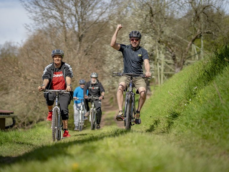Vier Männer fahren Fahrrad über eine Wiese und einer reckt die Faust in die Höhe.