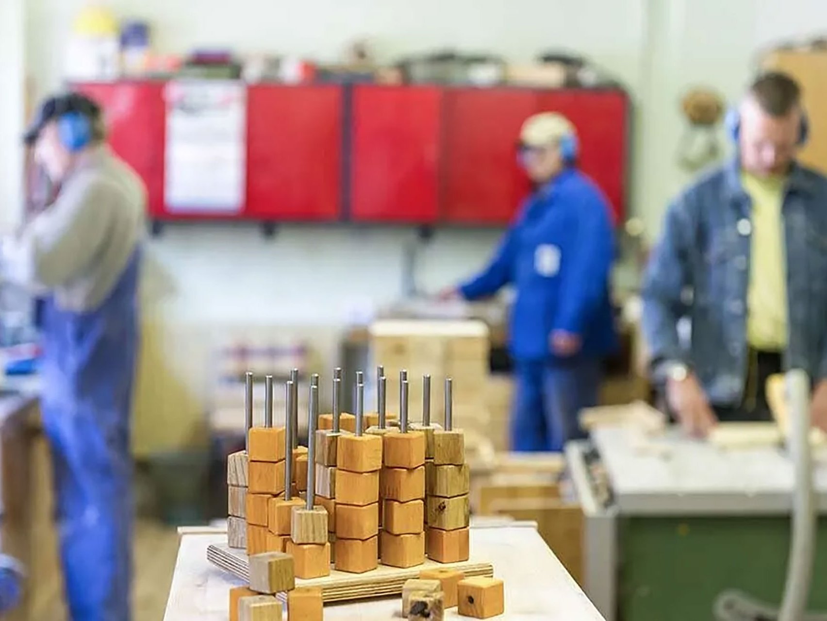 Werkstatt: Ein Kunstwerk aus Metall und Holzklötzchen - Im Hintergrund Arbeiter mit Gehörschutz beim Sägen und Bohren